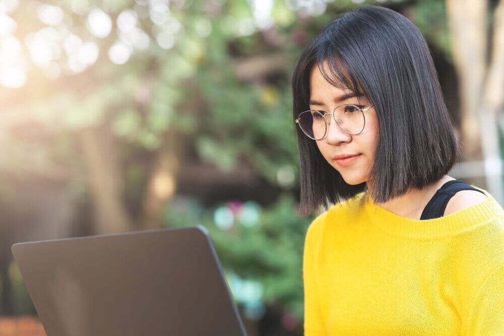 Young happy asian female student looking at computer laptop at outdoor cafe or coworking space. Young asian freelance stylist hipster, short hair and eyeglasses working outdoor concept.