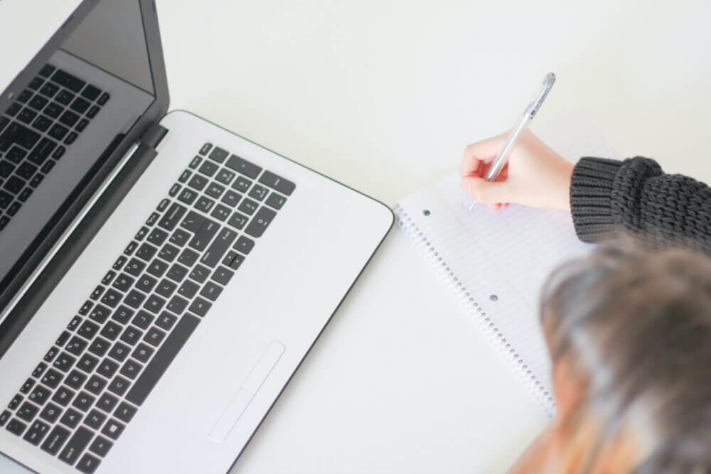 person writing notes next to Macbook