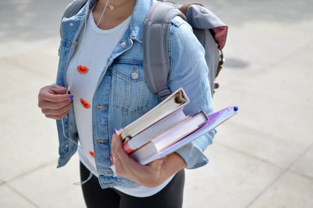 woman with denim jacket and backpack holding binder and textbooks in her hand