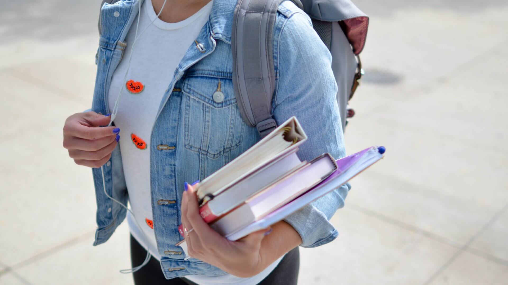 woman with denim jacket and backpack holding binder and textbooks in her hand