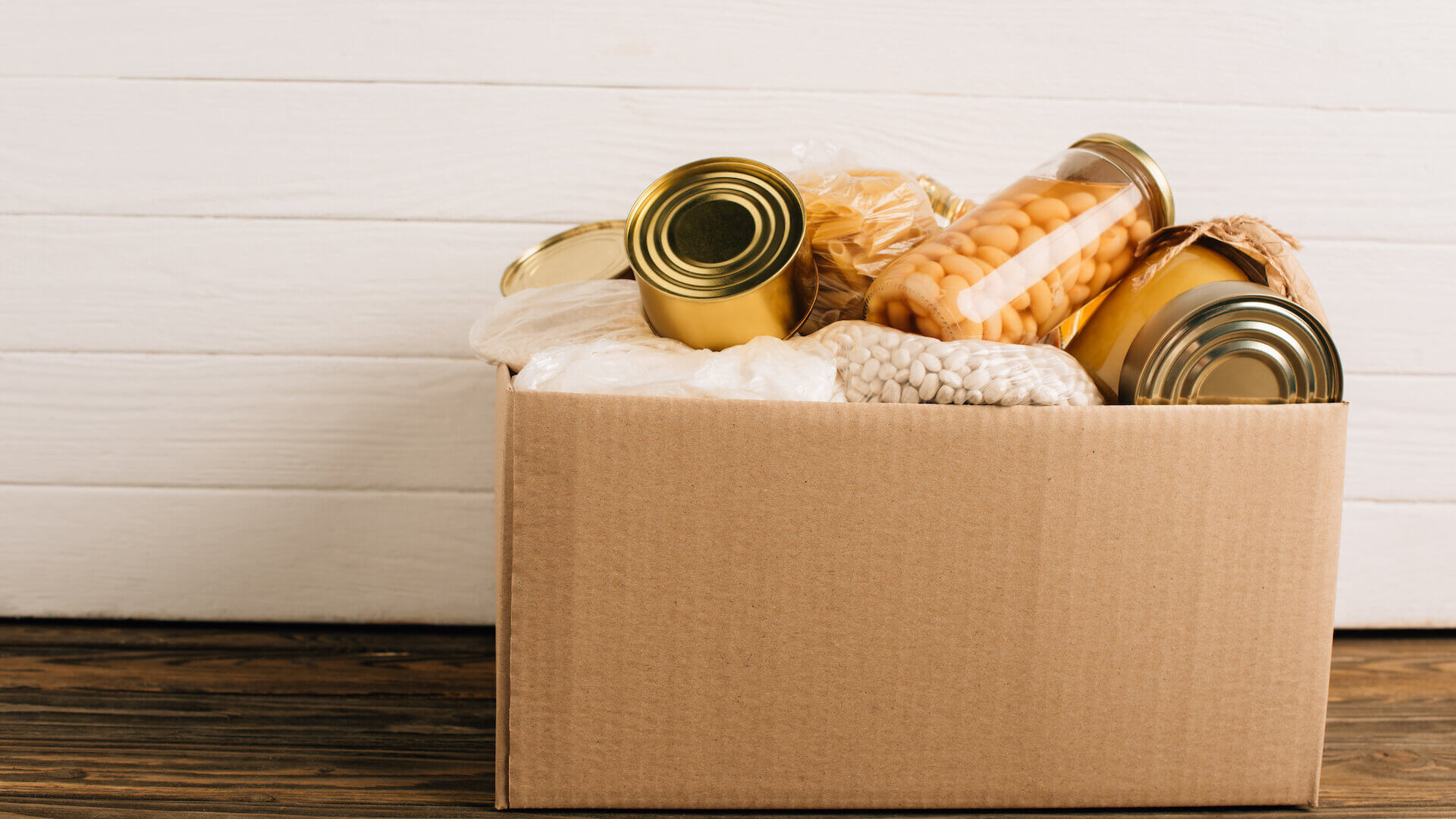 Cardboard box of canned food items on wooden shelf