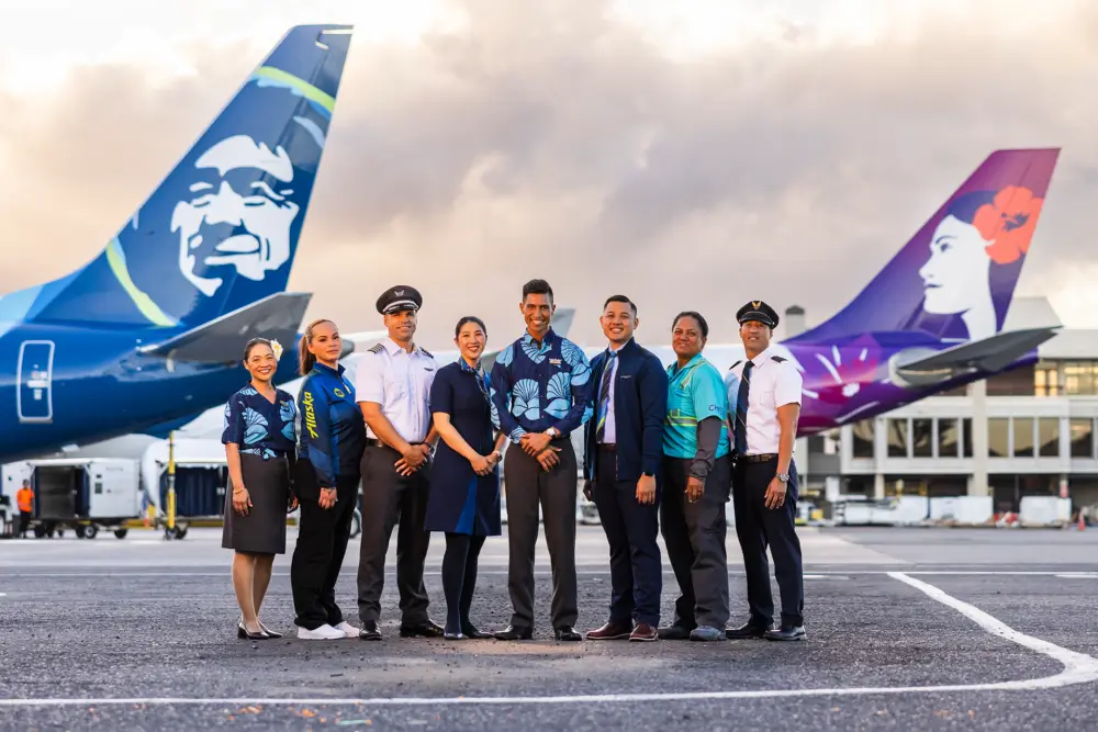 Alaska Airlines and Hawaiian Airlines employees stand in front of tail wings of their respective airplanes
