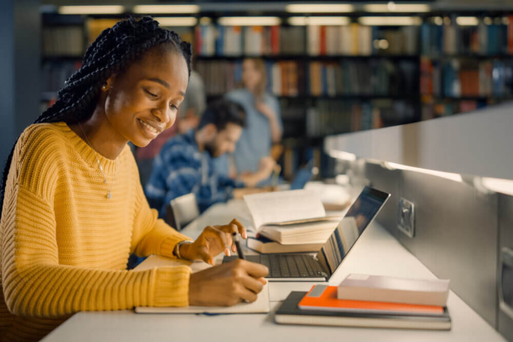 University Library: Gifted Black Girl uses Laptop, Writes Notes for the Paper, Essay, Study for Class Assignment. Diverse Multi-Ethnic Group of Students Learning, Studying for Exams, Talk in College