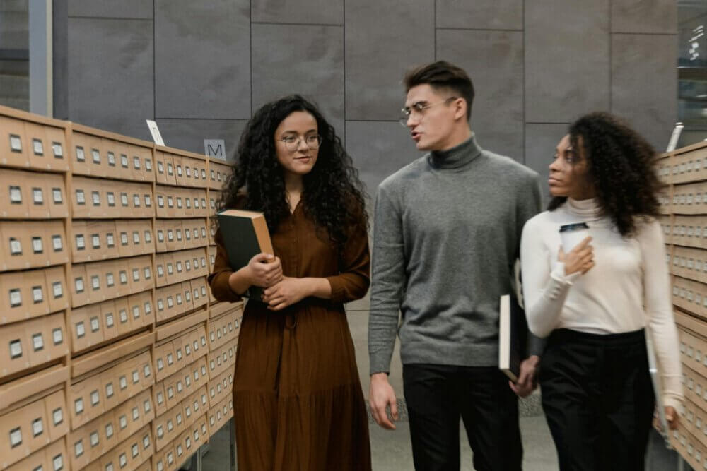 Three prospective students walking with books through the card catalogs of a library.