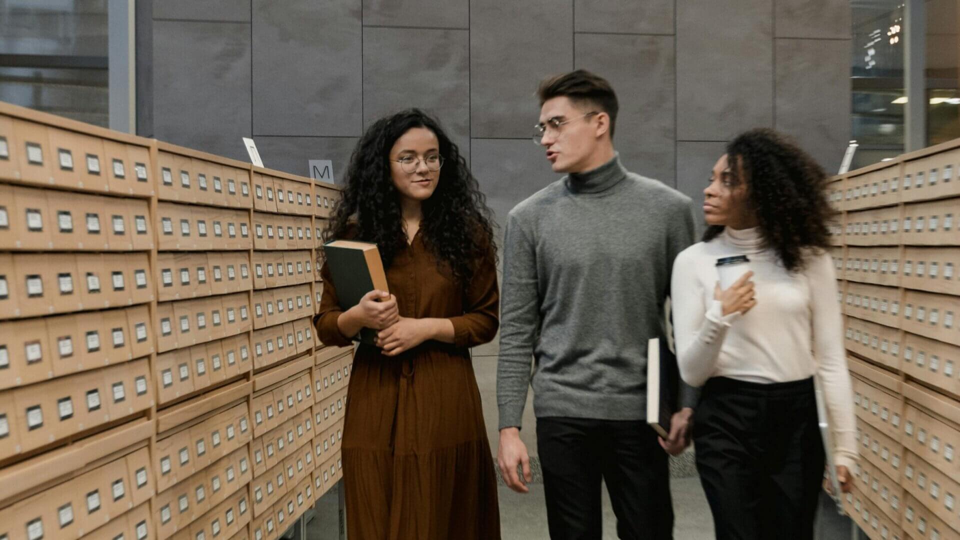 Three prospective students walking with books through the card catalogs of a library.
