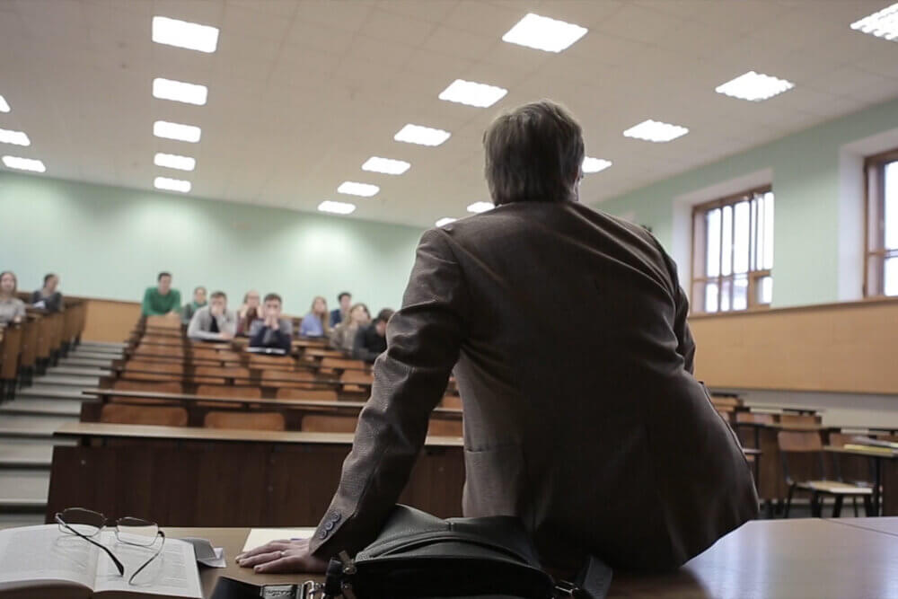 Back view of man presenting to students at a lecture theatre. View from the back of the teacher who is sitting on the table.