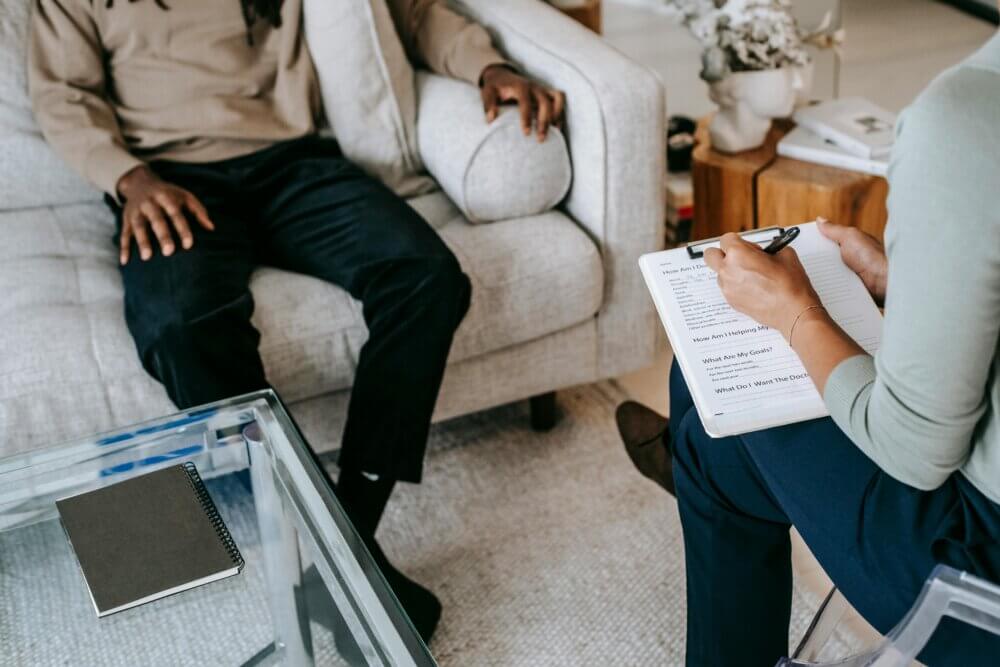 Male sitting on a couch, woman sitting across from the male in a chair writing notes down; during a therapy session.