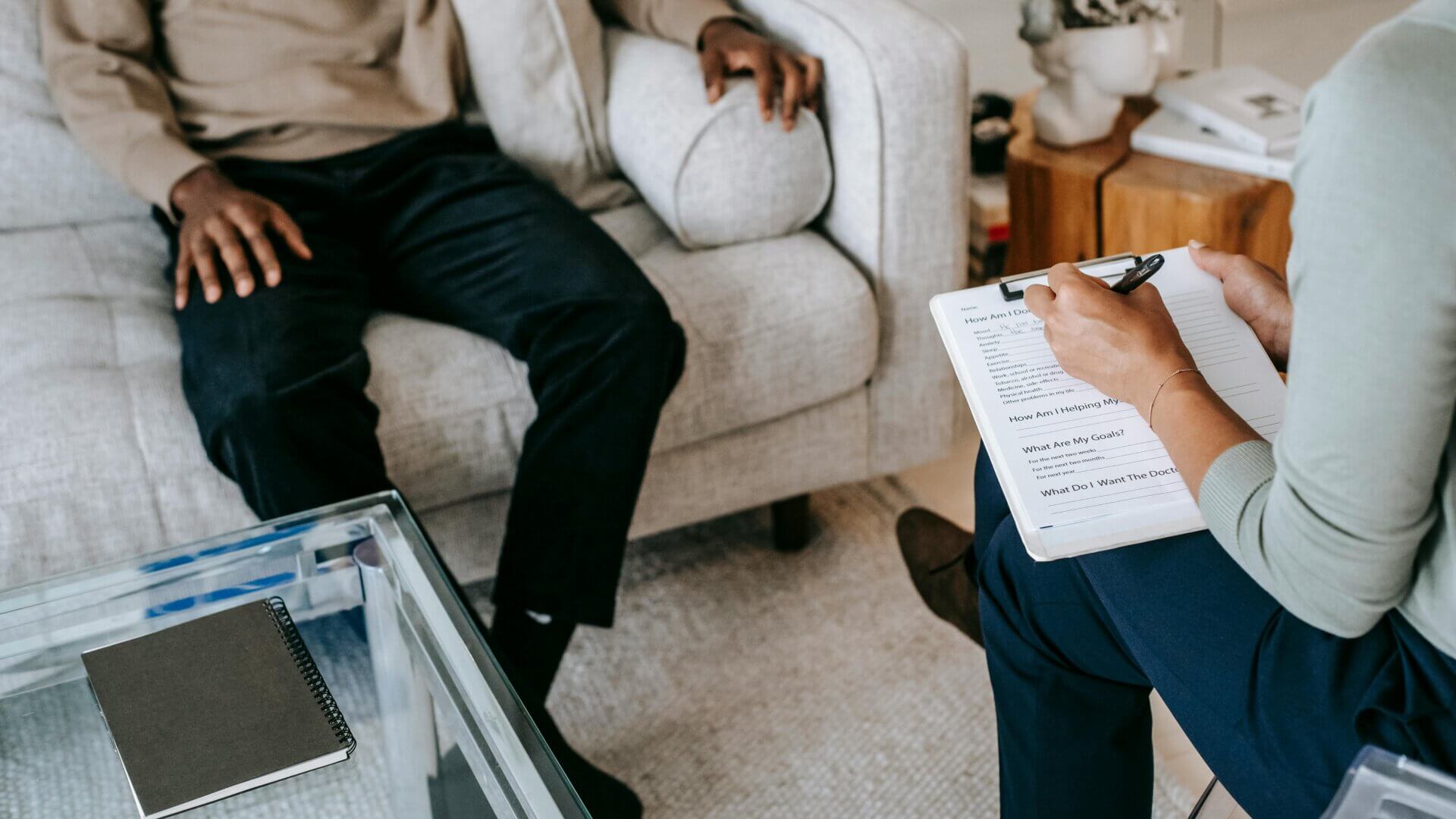Male sitting on a couch, woman sitting across from the male in a chair writing notes down; during a therapy session.