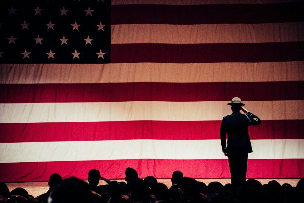 Military people saluting a large American flag with one person standing above the others.