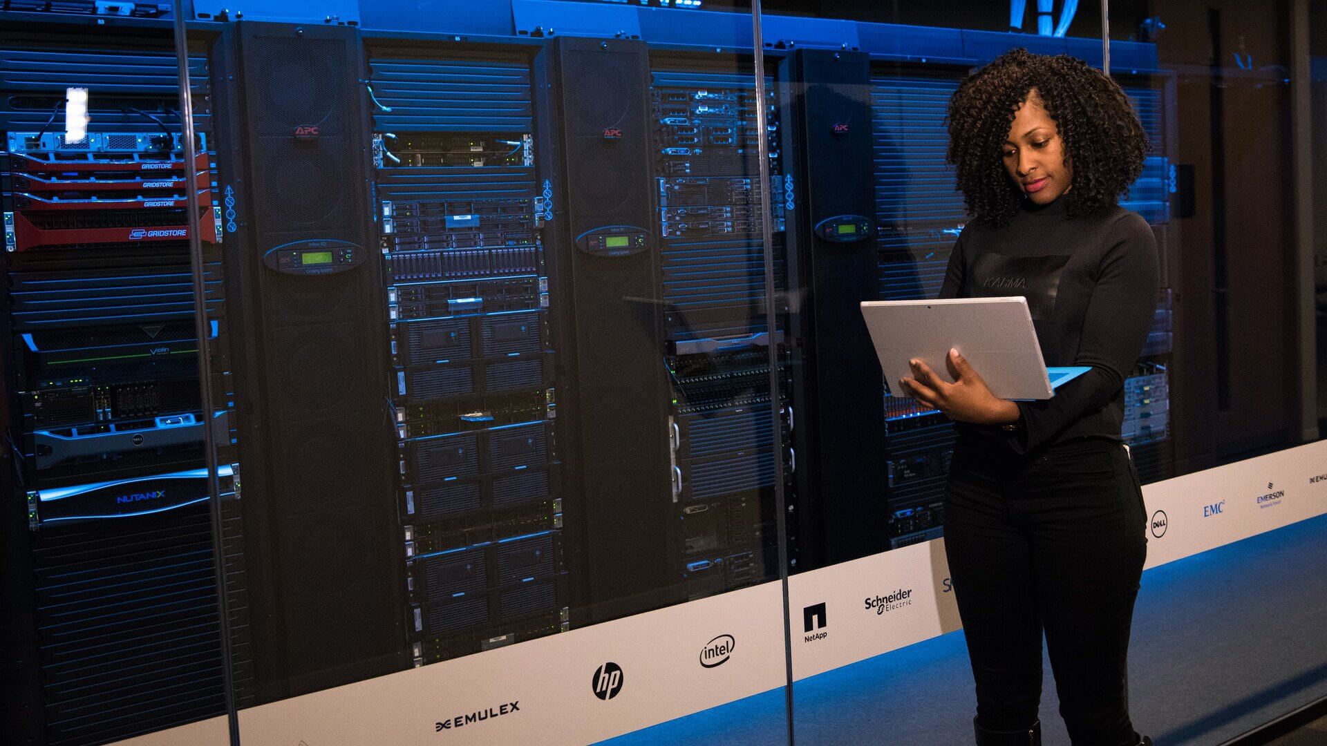 a woman standing with a laptop in front of a row of servers