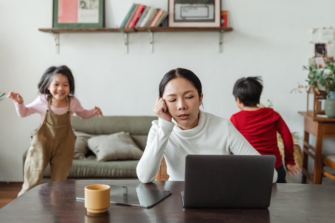 Mother appears stressed while working from home while her kids are playing in the background
