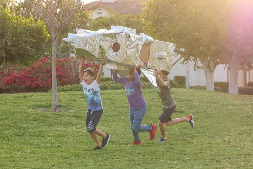 three boys running on grass with a homemade rocket ship