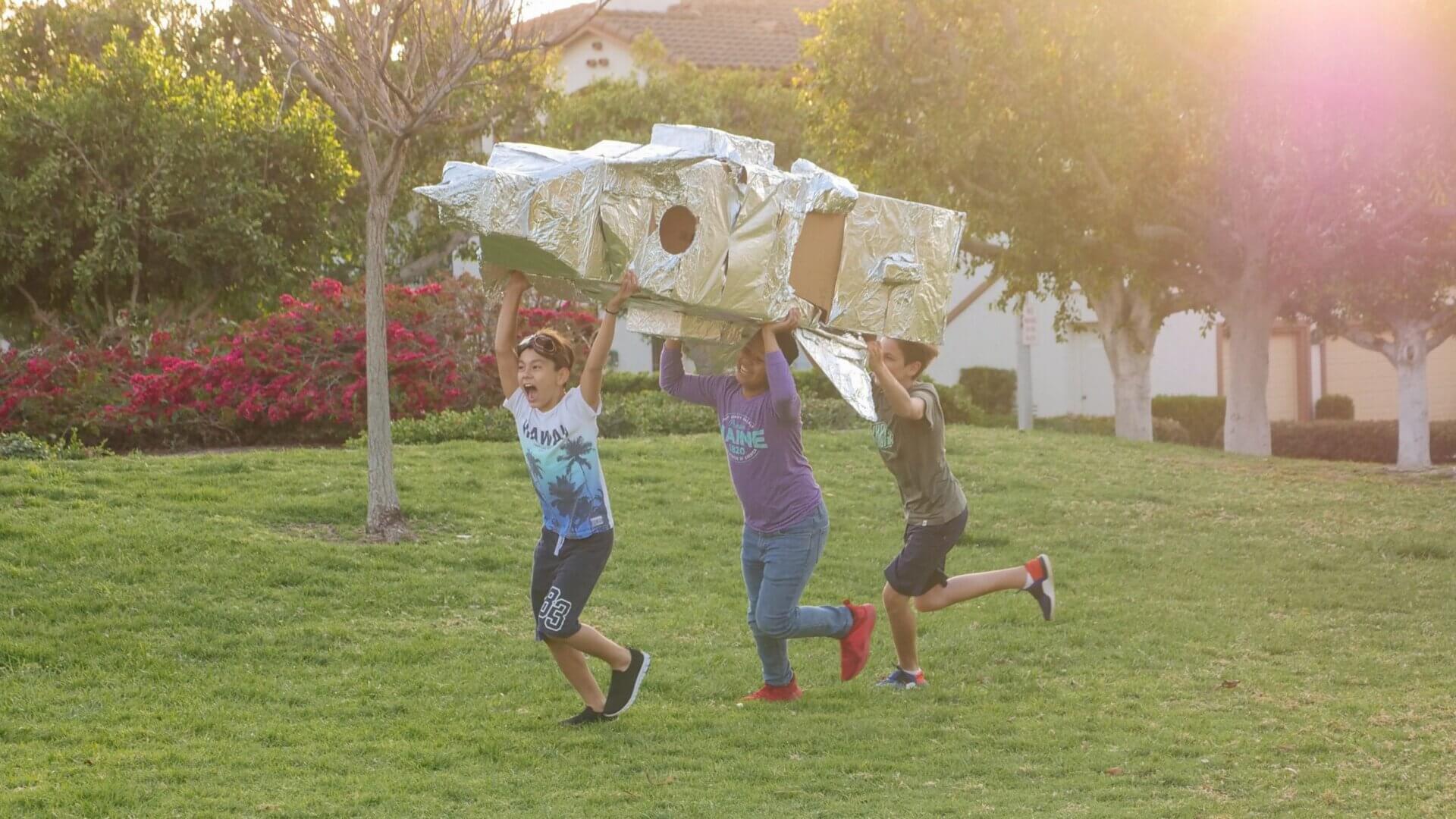 three boys running on grass with a homemade rocket ship