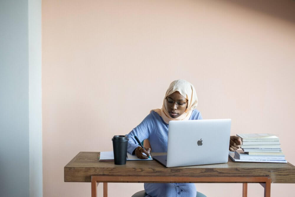 a woman writes in a notebook next to her open laptop