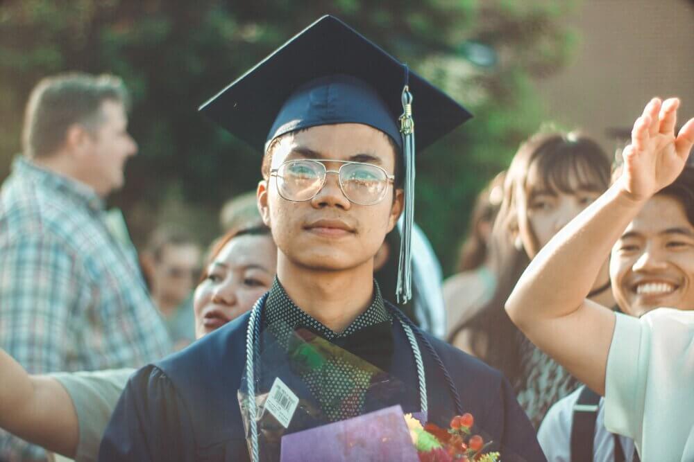 A boy looks at the camera holding his high school diploma, wearing a cap and gown