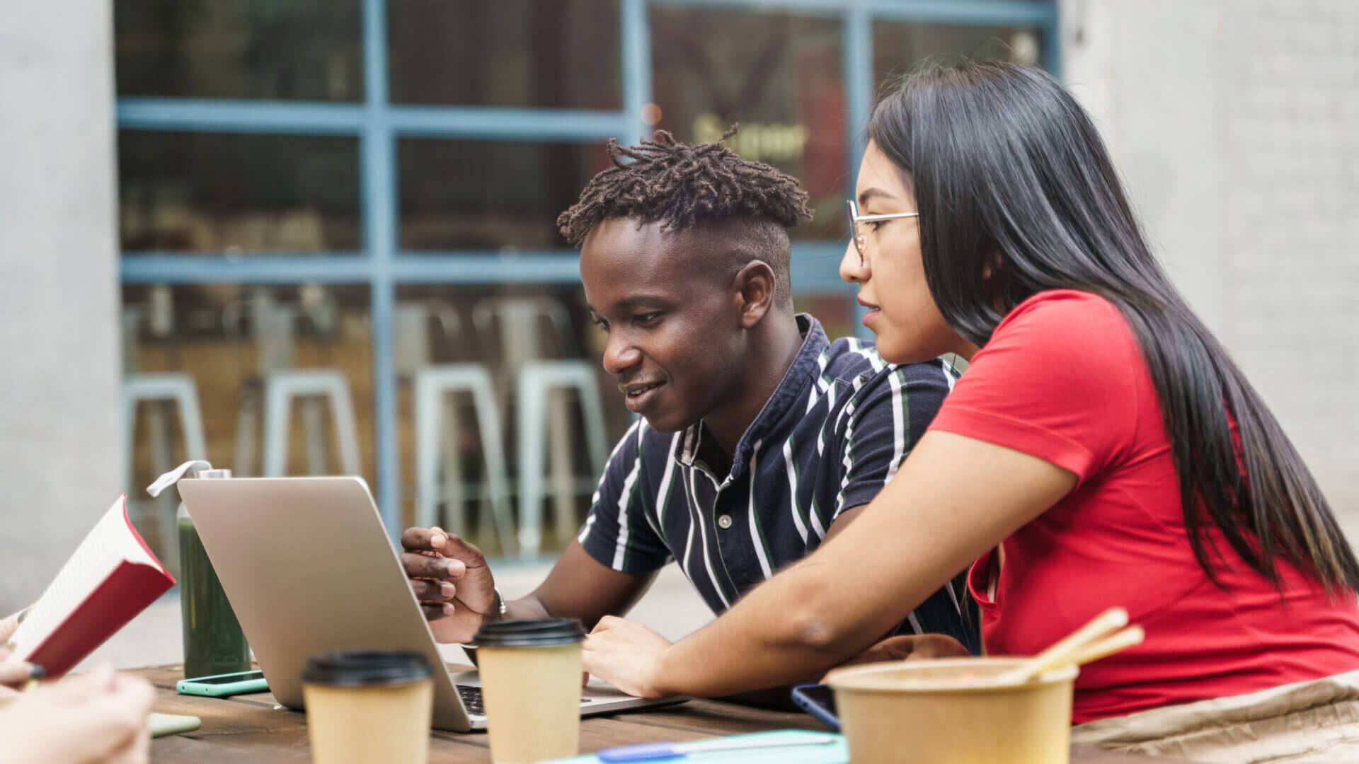 Two students working on a laptop while eating lunch and drinking coffee