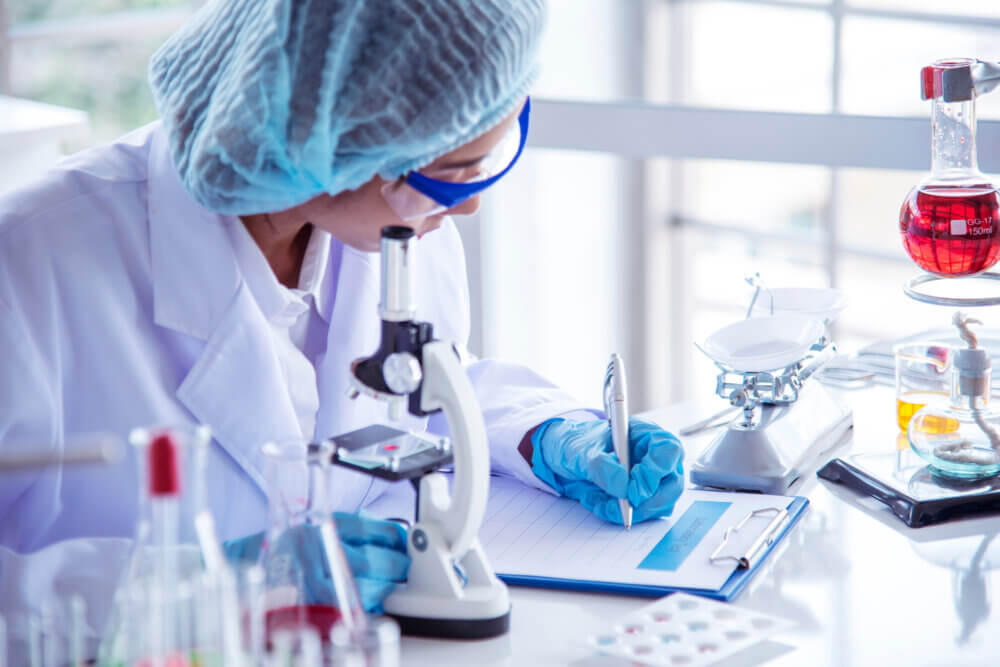 Scientist hand writes note information in medical lab looking at science microscope.