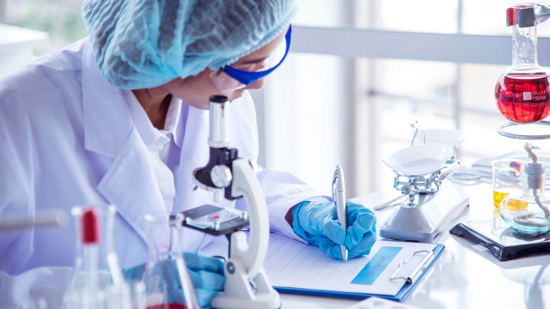 Scientist hand writes note information in medical lab looking at science microscope.